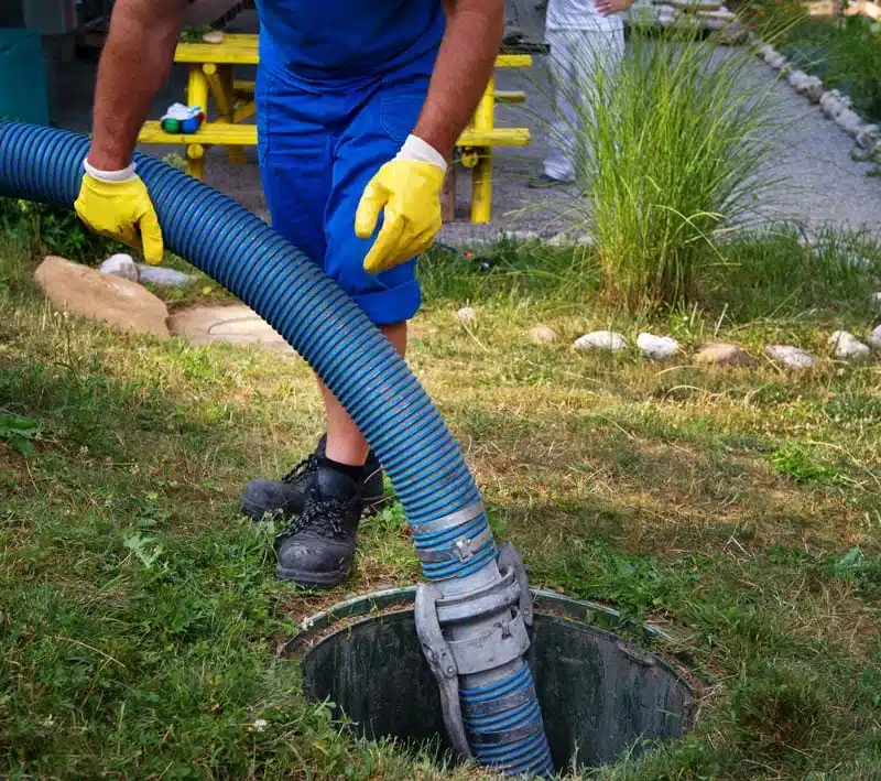 Worker lowering a large blue hose into an open tank access in a yard
