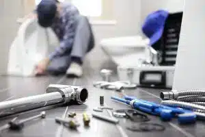 Plumbing tools and fittings laid out on a bathroom floor with a plumber working at a toilet behind them