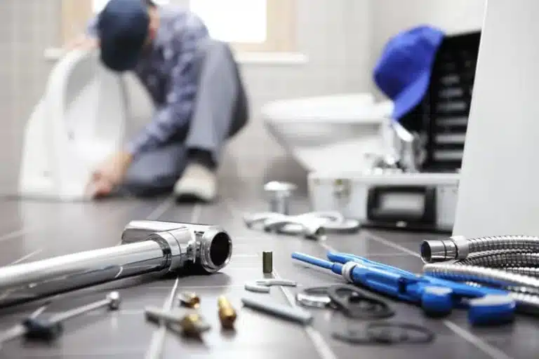 Plumbing tools and fittings laid out on a bathroom floor with a plumber working at a toilet behind them
