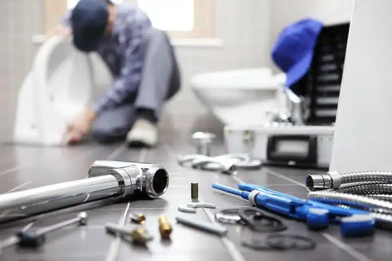 Plumbing tools and fittings laid out on a bathroom floor with a plumber working at a toilet behind them