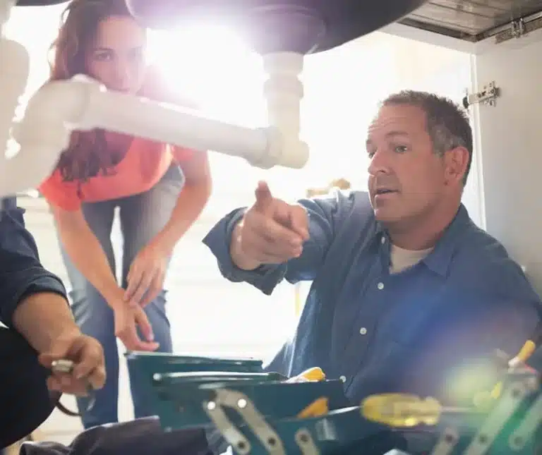 Plumber under a kitchen sink explaining a repair to a homeowner