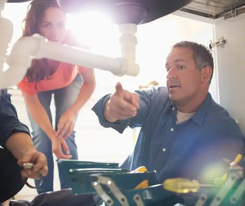 Plumber under a kitchen sink explaining a repair to a homeowner