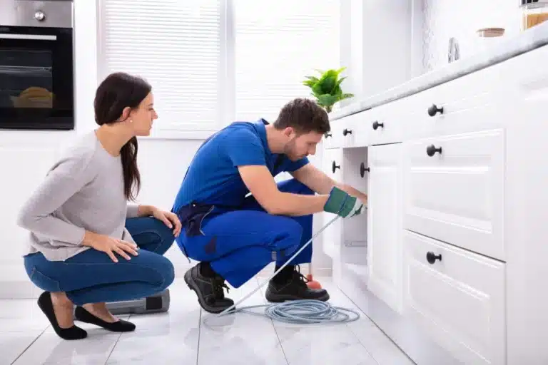 Plumber running a drain cable under a kitchen sink while the homeowner watches
